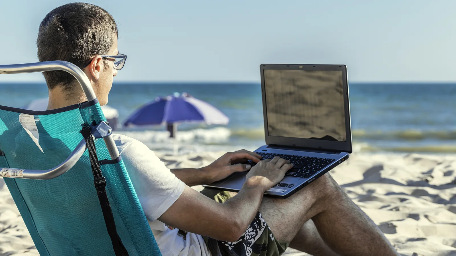 Mężczyzna pracujący na laptopie z leżaka na plaży nad morzem.